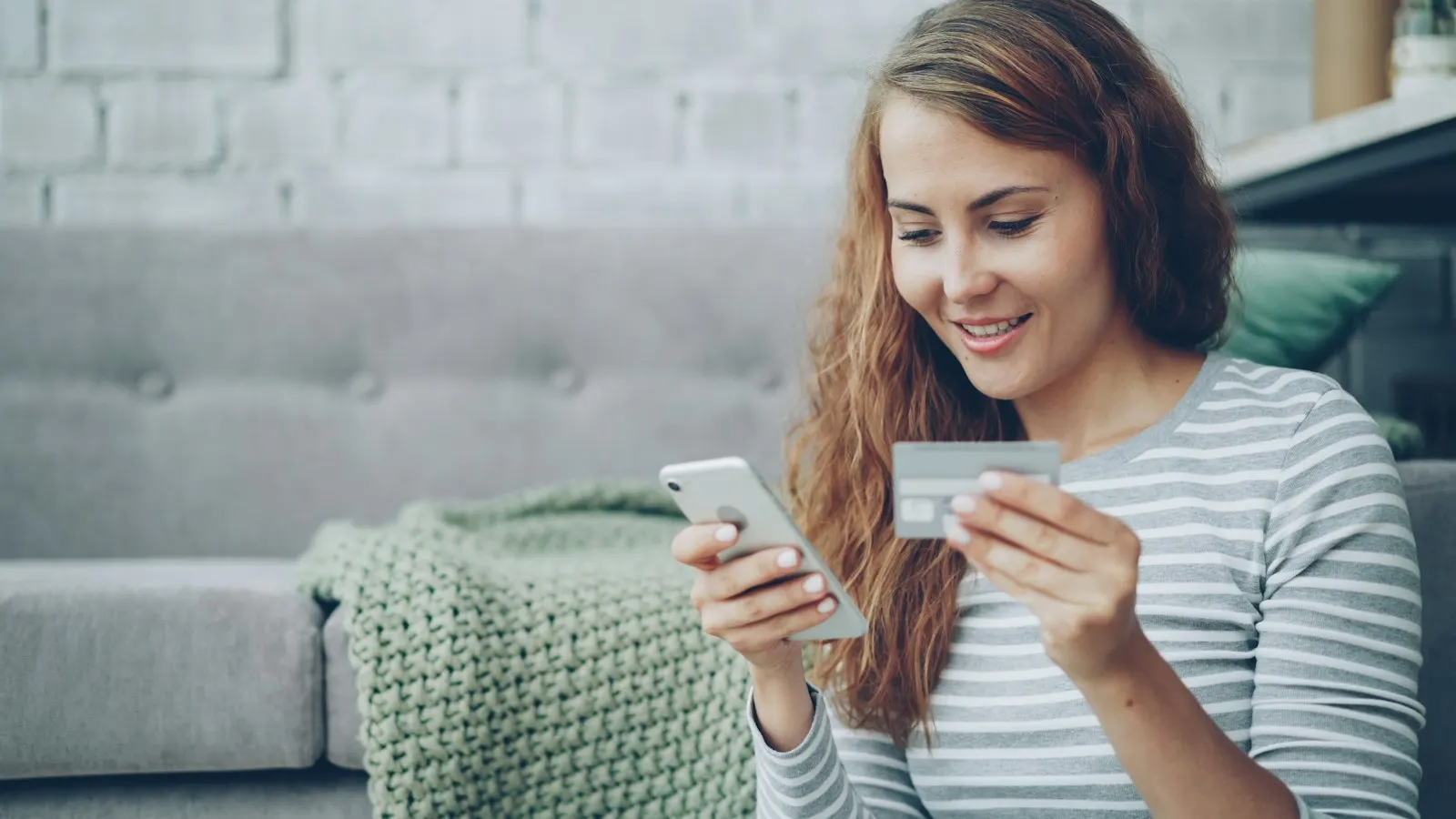 A woman at home smiling as she pays with her card on her phone